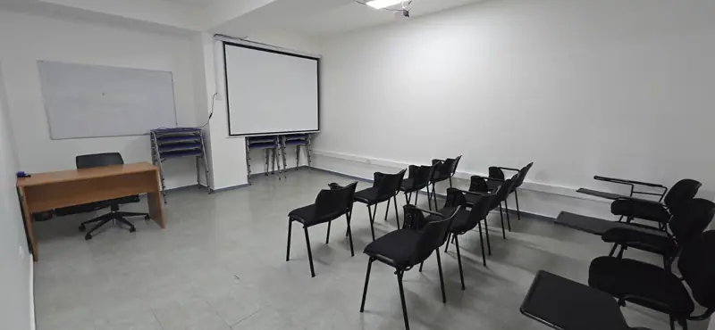 Sala de clases con pizarra, escritorio y sillas con paleta.
