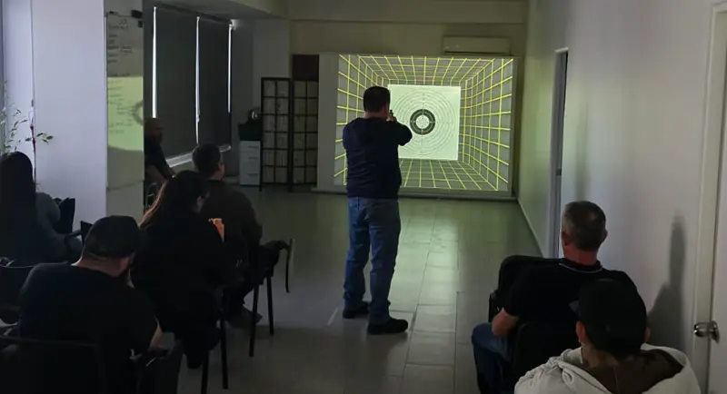 Alumnos sentados observan a un compañero que practica frente a la diana proyectada del simulador.