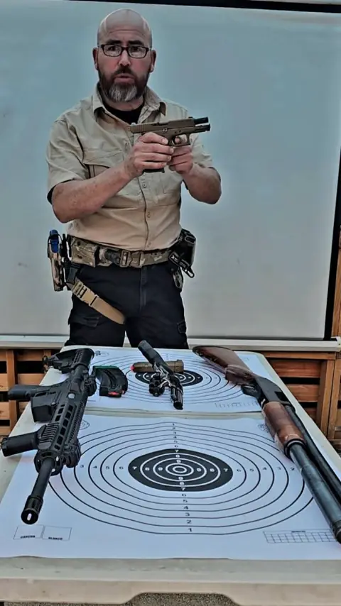 Instructor muestra una pistola; sobre la mesa hay otros fusiles, una escopeta y dianas.