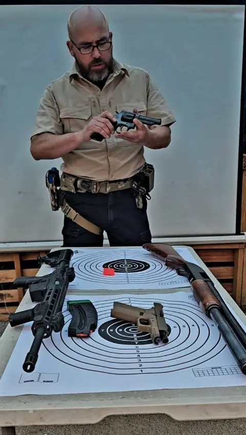 Instructor muestra un revólver frente a la mesa con armas largas y dianas.