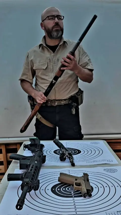 Instructor sostiene una escopeta frente a la mesa con armas y dianas.
