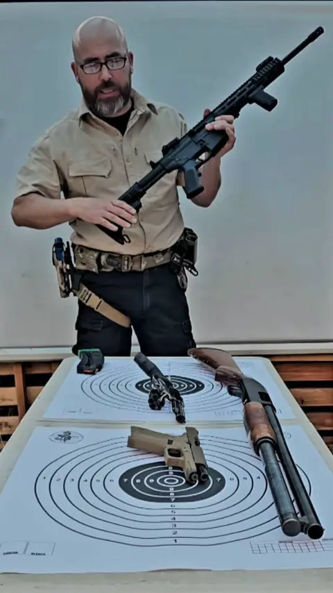 Instructor sostiene un rifle frente a la mesa con armas y dianas.