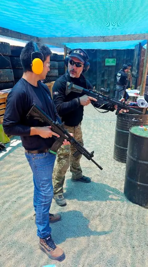 Alumno con fusil y protección auditiva junto a un instructor en el polígono exterior.